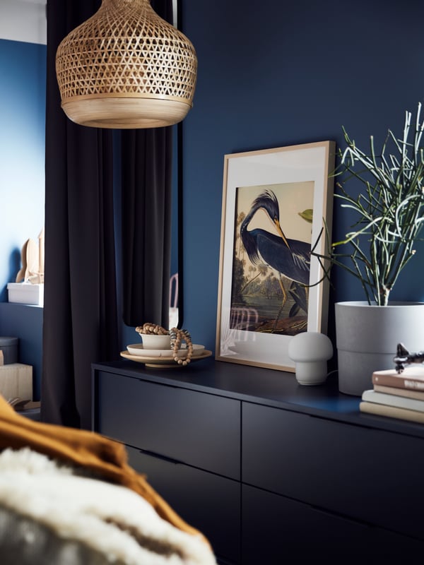 A black-blue NORDMELA chest of drawers with a framed picture, pile of books and plant on top, plus a bamboo pendant above.