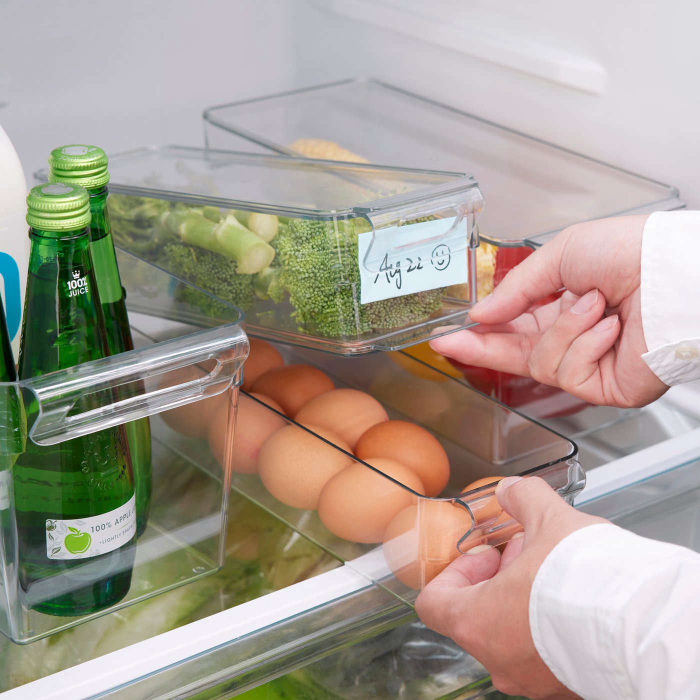 Hands pulling out a KLIPPKAKTUS storage box in the fridge, filled with eggs, broccoli, and drinks.