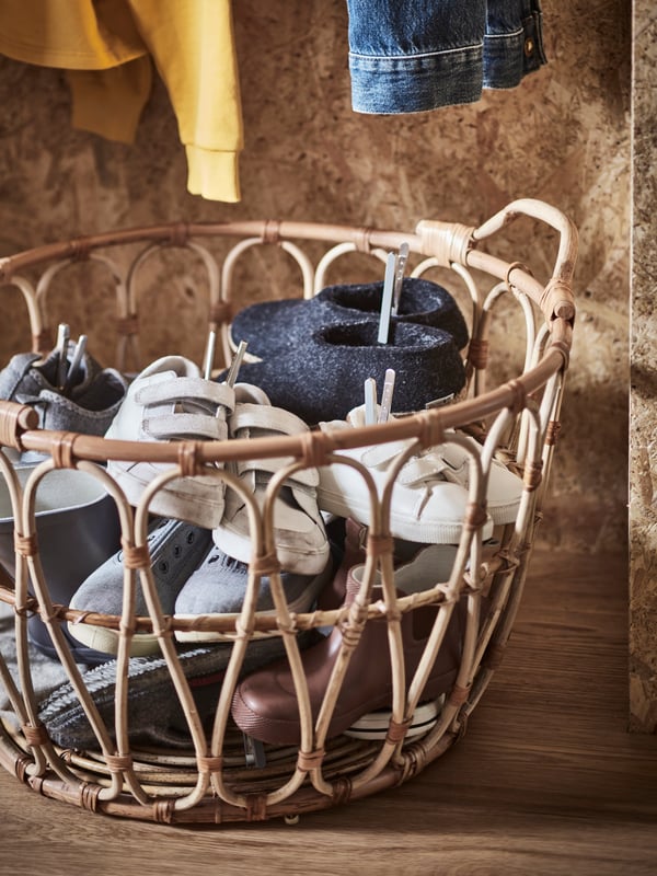 A SNIDAD rattan basket with shoes inside standing on the floor of a hallway below various hanging clothes.