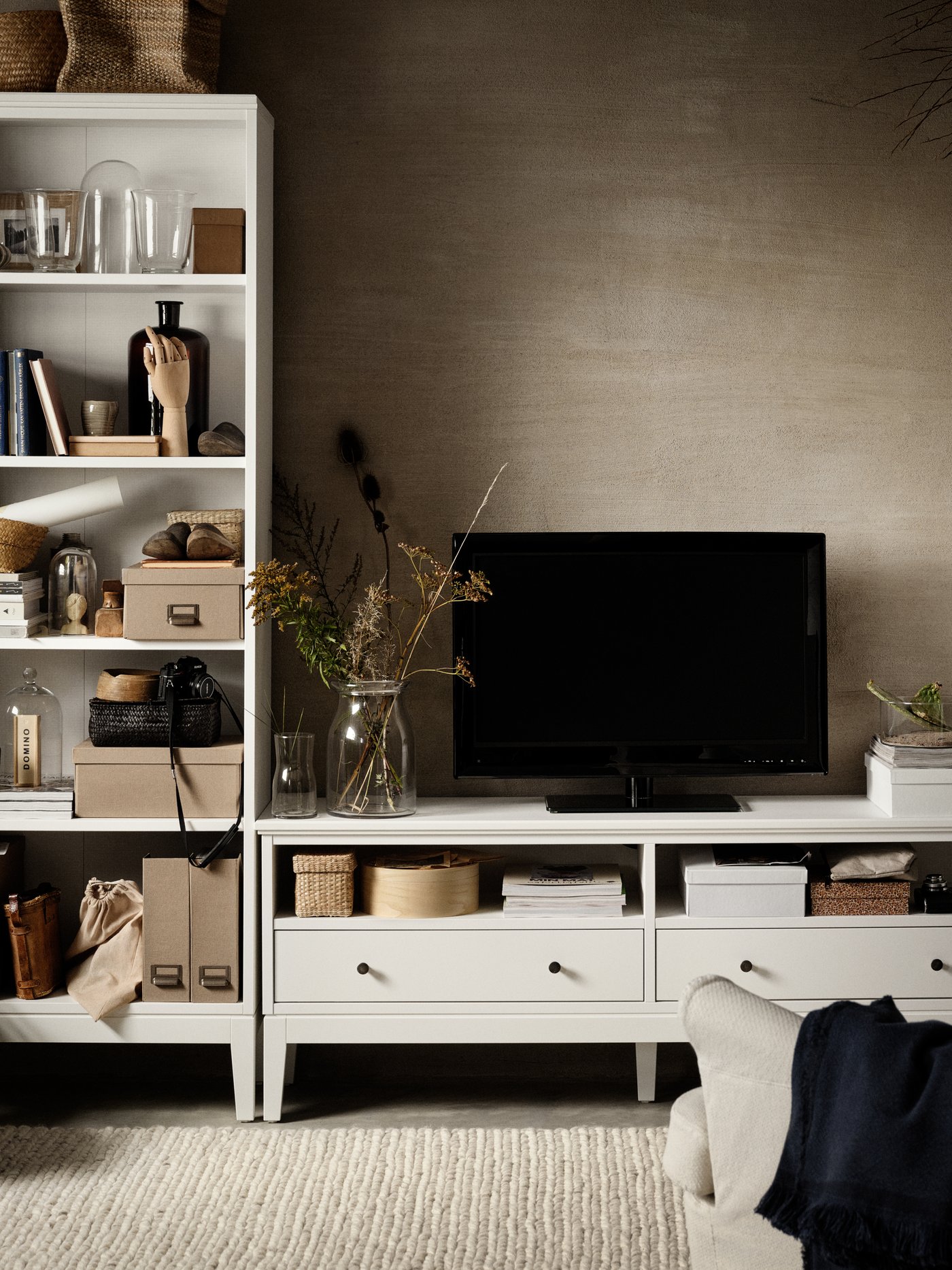 A white IDANÄS bookcase with boxes, magazine files and decorations. Beside it stands a TV on a white IDANÄS TV bench.