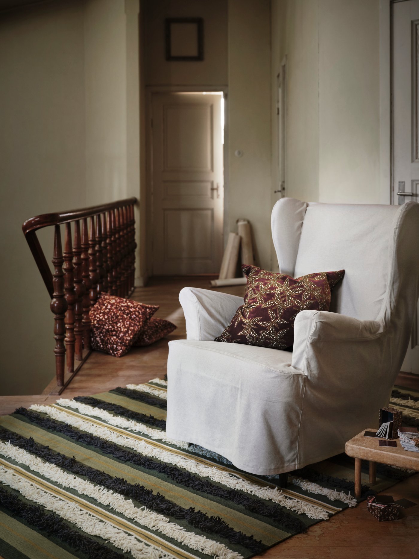 An armchair with a beige STRANDMON armchair cover and a red and gold pillow stand on a rug at the top of some stairs.