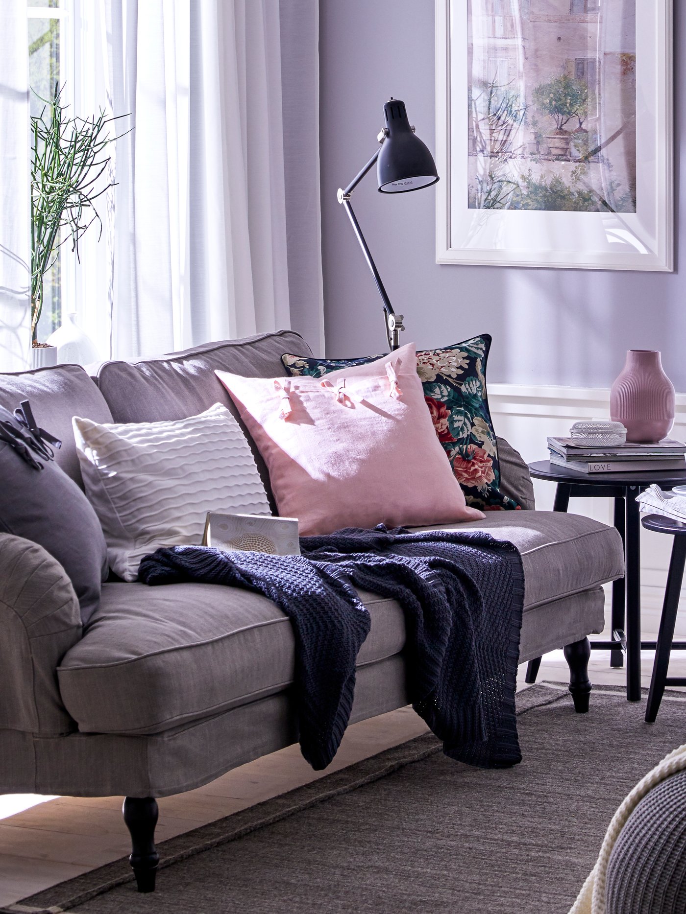 Ein Plaid und mehrere Kissen, u. a. mit einem hellrosafarbenen AINA Kissenbezug liegen auf einem Sofa nahe eines Fensters.