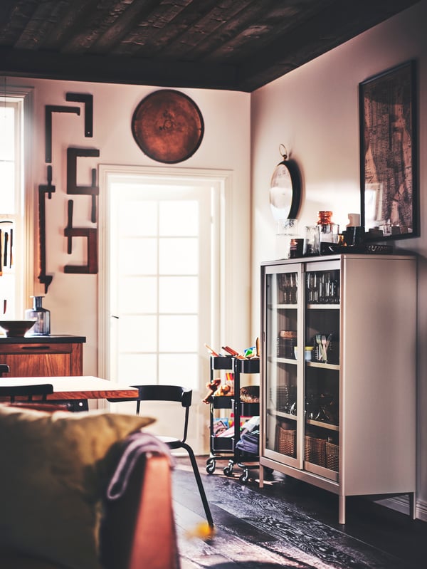 A dining area with a YNGVAR chair, a beige IDÅSEN cabinet with sliding glass doors and two black RÅSKOG trolleys.