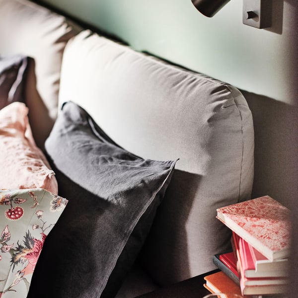 The head of a SAGESUND upholstered bed in Diseröd brown with cushions and pillows in various colours on it, and a lamp above.