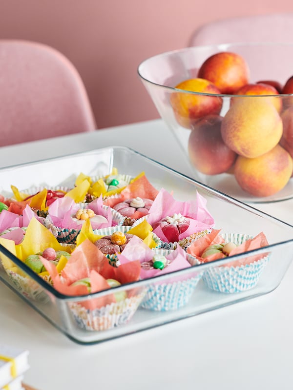 A flat glass serving dish filled with colourful cakes, beside a deeper glass dish full of fruit on a white table top.