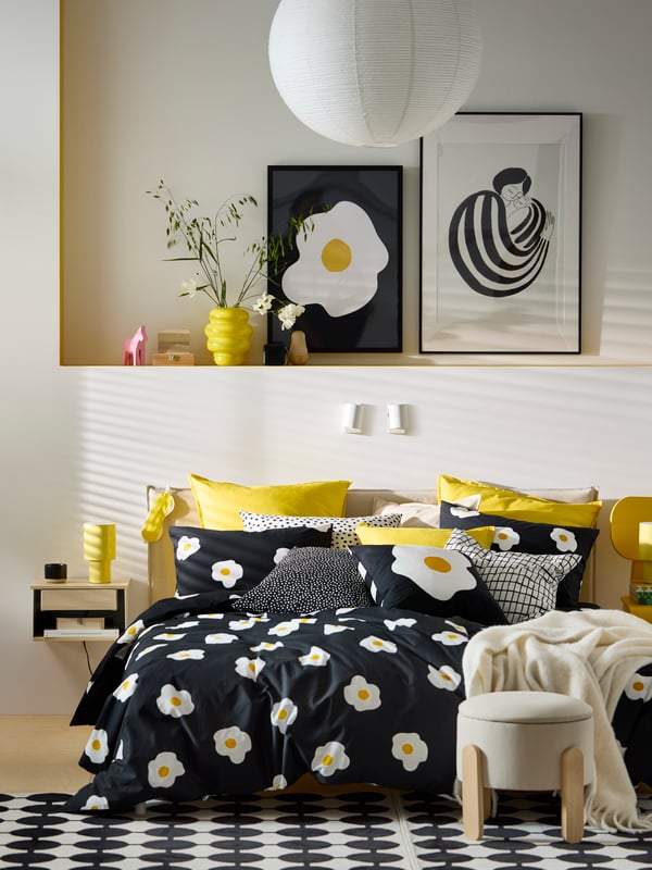 Black, white and yellow bedroom with white walls and fried-egg themed bedding. Two framed prints and yellow vase above bed.