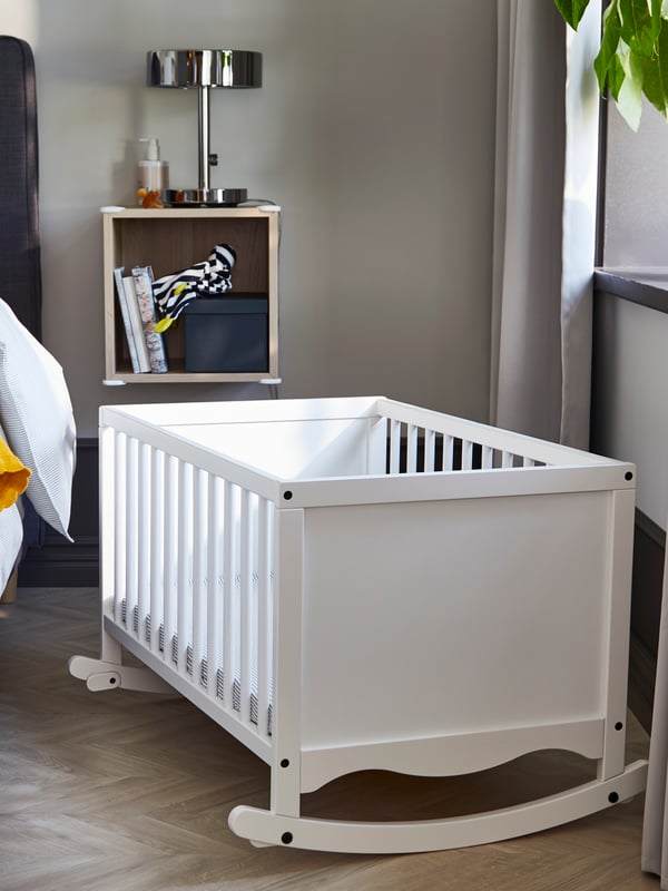 A white SOLGUL cradle sits in a bedroom near a window and an EKET wall cabinet with a STOCKHOLM table lamp on top.