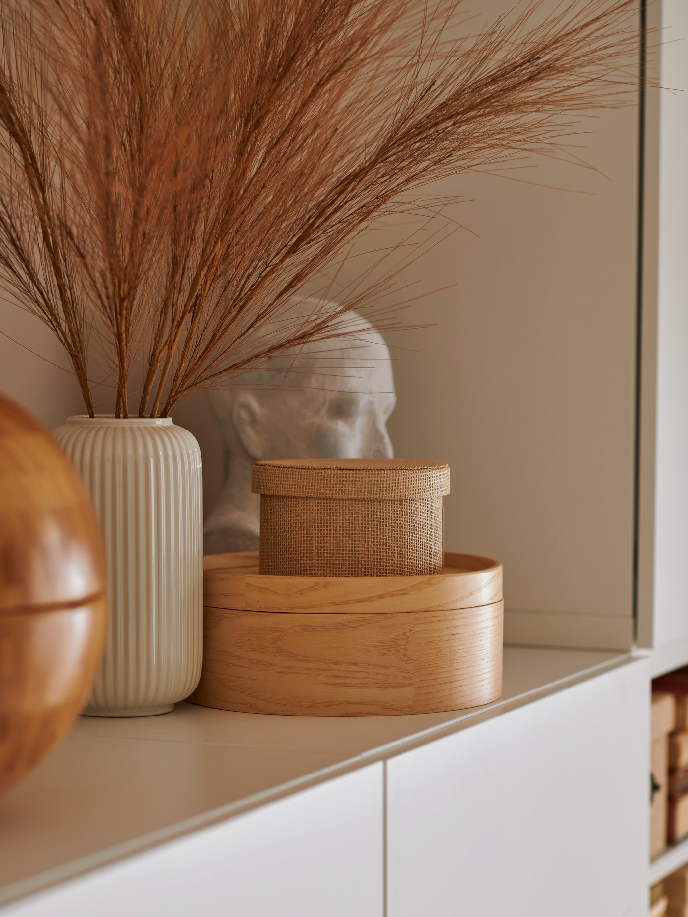 Auf einem minimalistischen weißen Regal stehen eine gerippte Vase mit getrockneten braunen Gräsern, Holzkisten und eine abstrakte Skulptur. Die Szene vermittelt ein ruhiges, natürliches Gefühl.