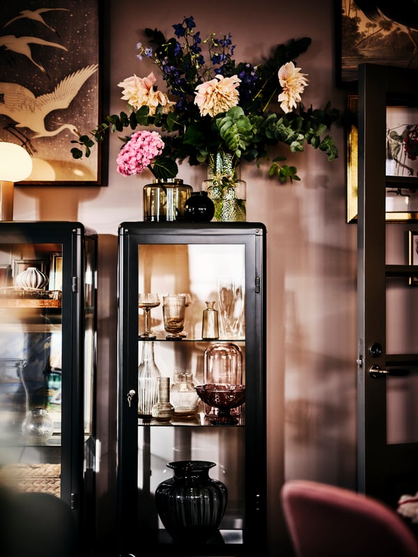 Two FABRIKÖR glass-door cabinets filled with glassware, one with two vases of fresh flowers on top, the other with a lamp.