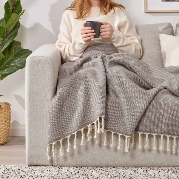 A person sitting on VIMLE light grey two seater sofa, holding a dark grey DINERA mug and covering their legs with a ULLTISTEL grey throw in the living room.