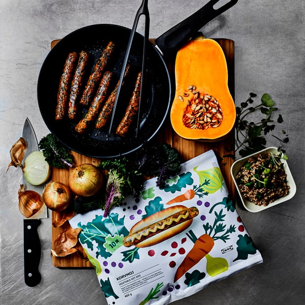 Hier ist ein detaillierter ALT‑Text unter 150 Zeichen, ohne Erwähnung eines Gesichts:
Ingrédients et saucisses végétales dans une poêle, entourés de courge, d’oignons, d’herbes et d’un emballage alimentaire coloré.