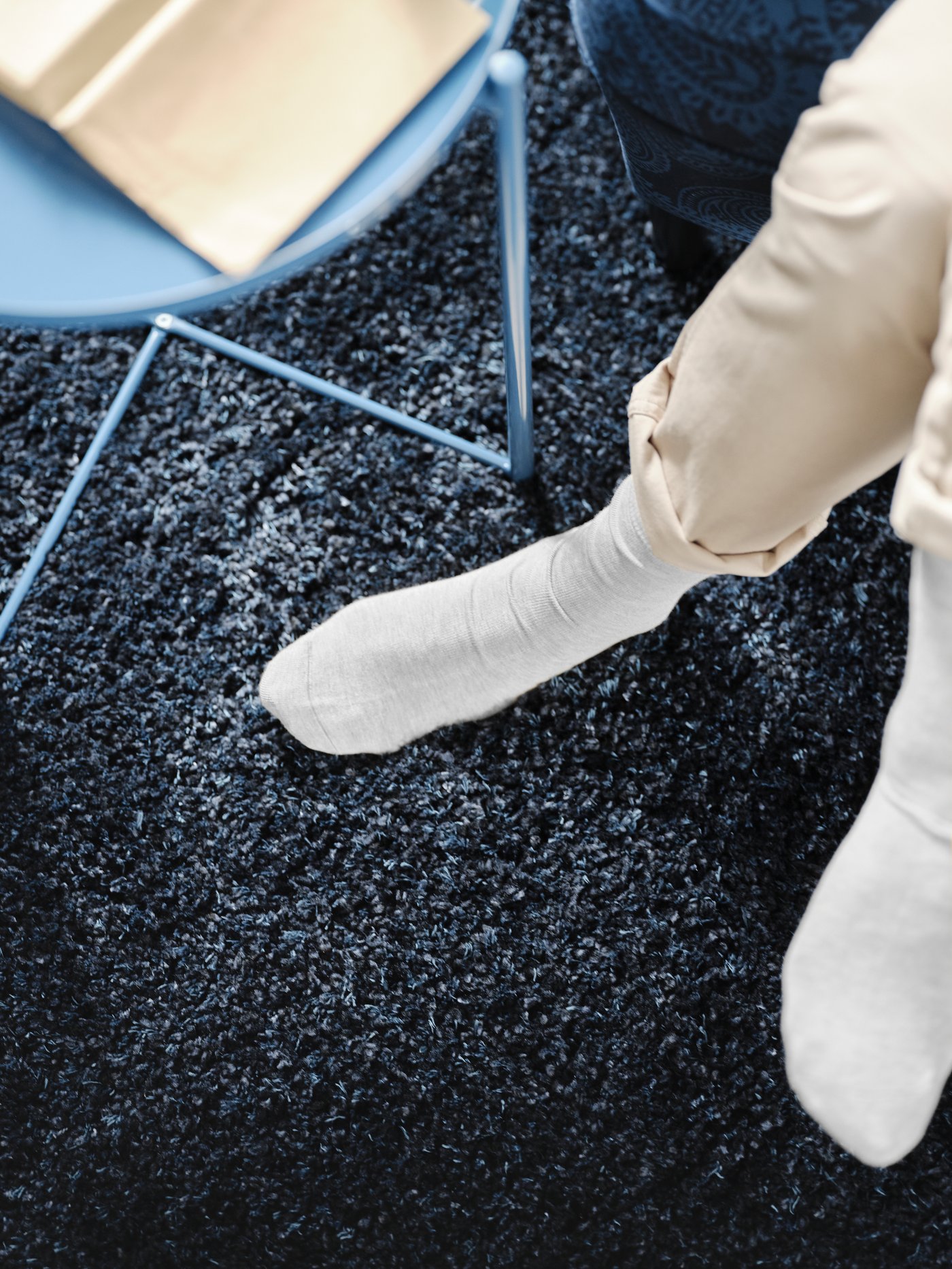 A dark blue VOLLERSLEV rug with high pile, a round, blue tray table holding a book, and a pair of feet with white socks.