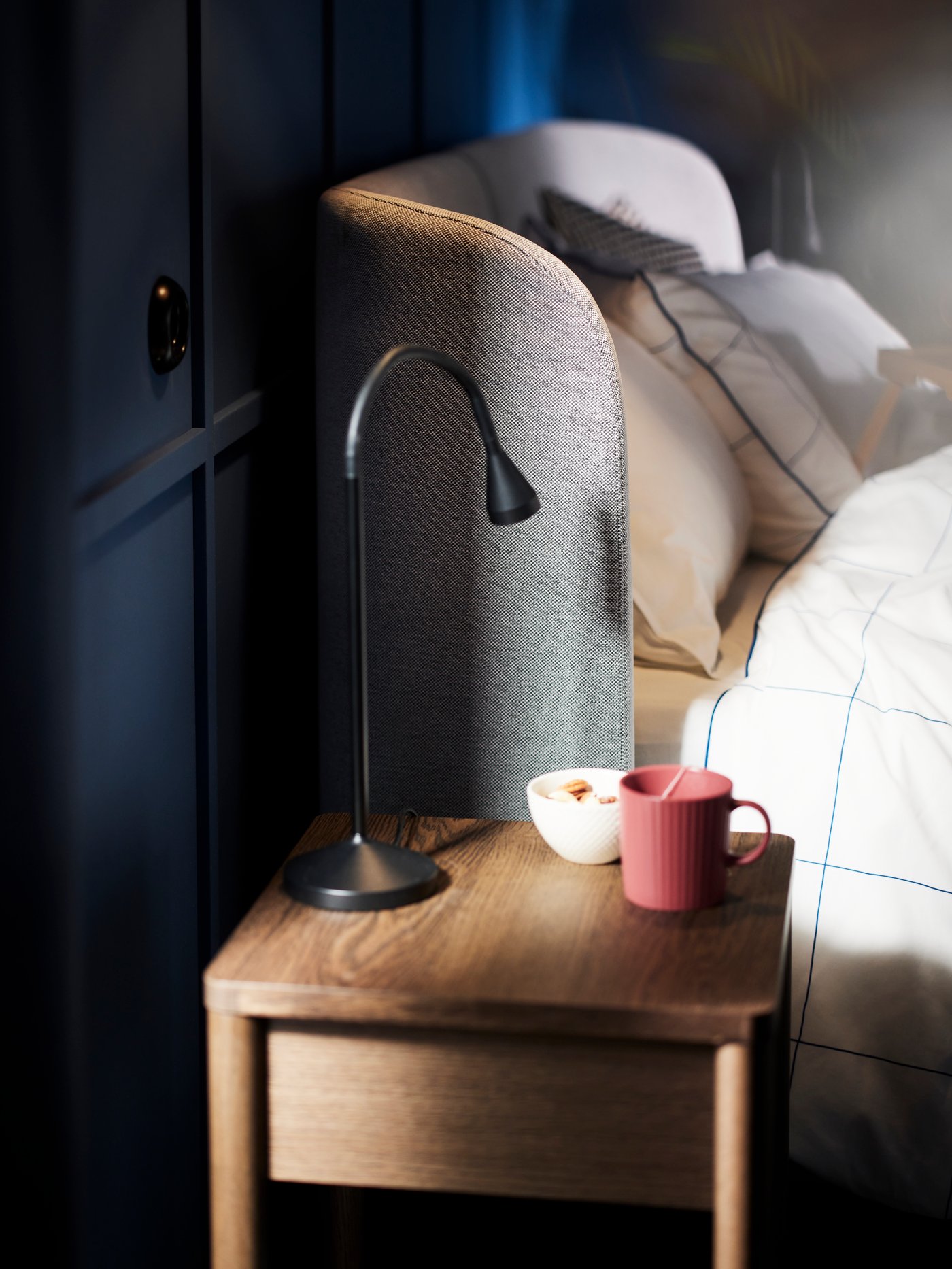 The head of a TUFJORD upholstered bed with a TONSTAD bedside table beside it which has a black NÄVLINGE LED work lamp on top.