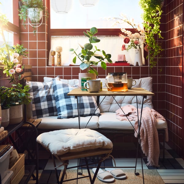 A cosy balcony features a black TÄRNÖ outdoor table, surrounded by plants, cushions, and a glass pot with tea.