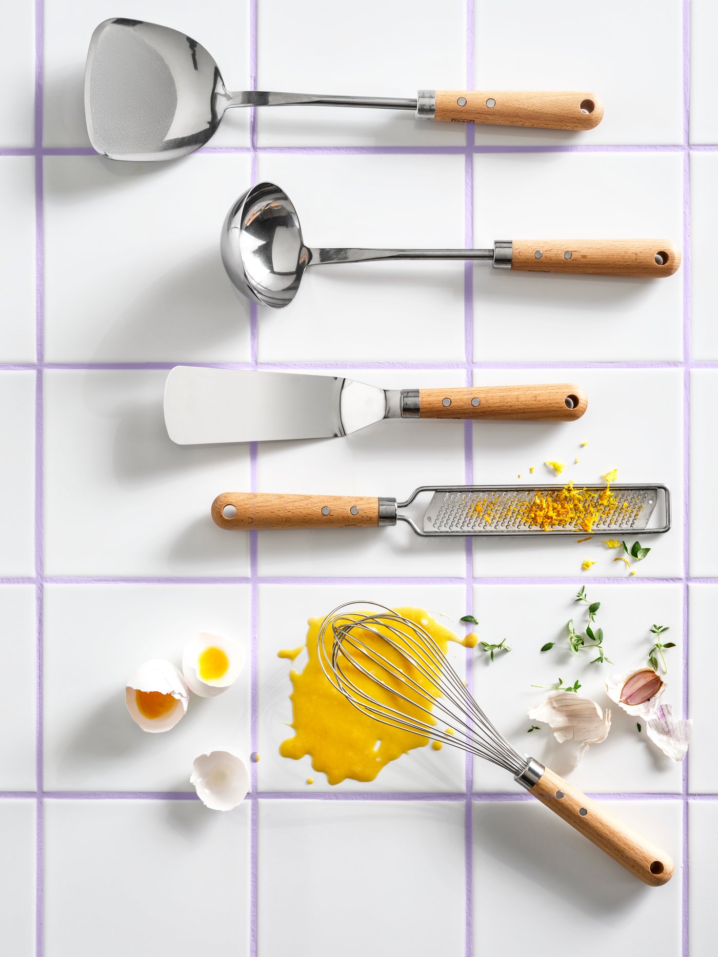 A top view of different VARDAGEN cooking utensils, placed on a white tiled surface together with eggs, garlic and herbs.