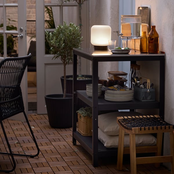 A black GRILLSKÄR outdoor kitchen island shelf unit with plates, cutlery, food, and an IKEA 365+ jar with tap on a patio.