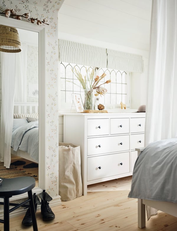 A white HEMNES chest with 8 drawers in front of a window, with a vase of dried flowers on it. A mirror is on the wall nearby.