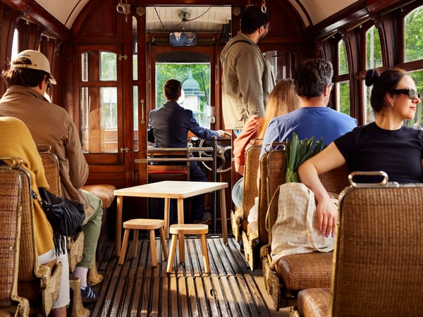 A small wooden table sits in the aisle of a tram, surrounded by passengers enjoying a sunny ride in wicker seats.