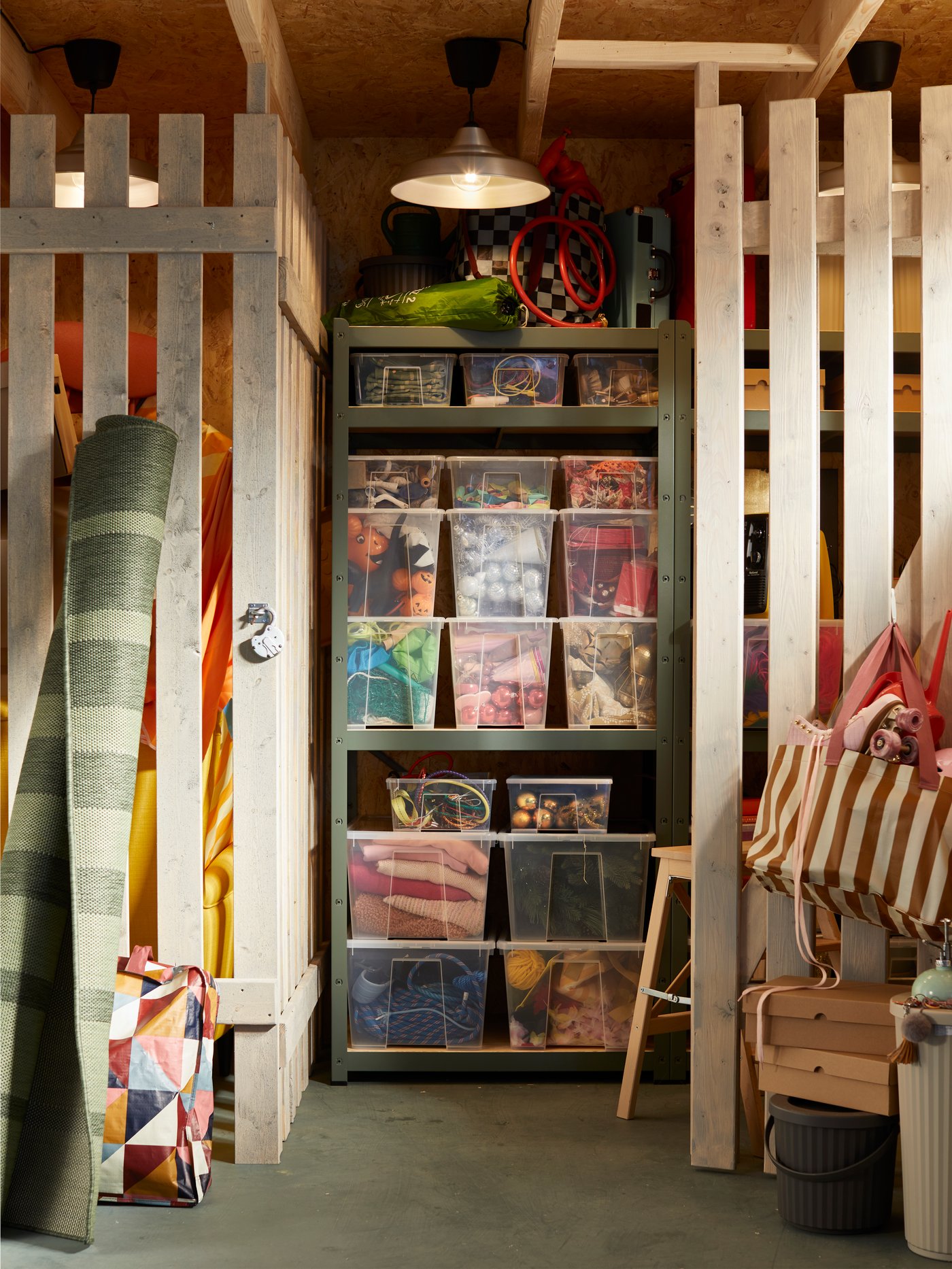 Grote IKEA-opbergnis met een hoge metalen kast vol stapelbare doorzichtige SAMLA-opbergboxen, een HEKTAR-hanglamp erboven, opgerolde vloerkleden tegen houten lattenwanden en kleurrijke tassen en accessoires op rekken.