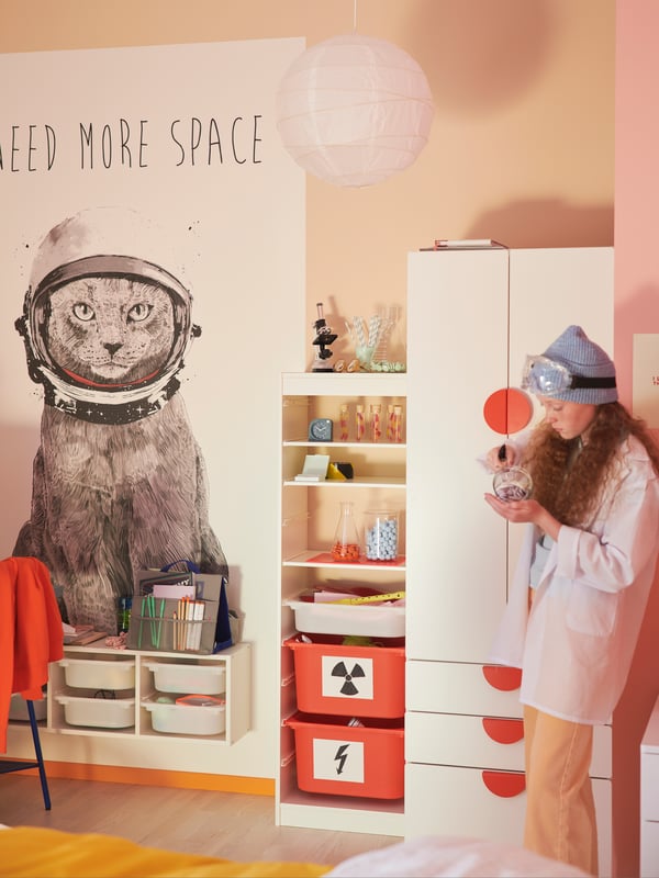 A girl holding a chemistry beaker stands by a white SMÅSTAD wardrobe with drawers, in a corner of a pastel-colours room.