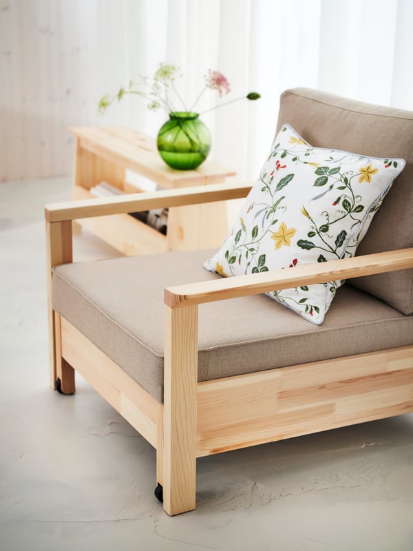A floral cushion lies on a beige and wood TORNSBORG armchair-bed near a bench in a calm white living room.