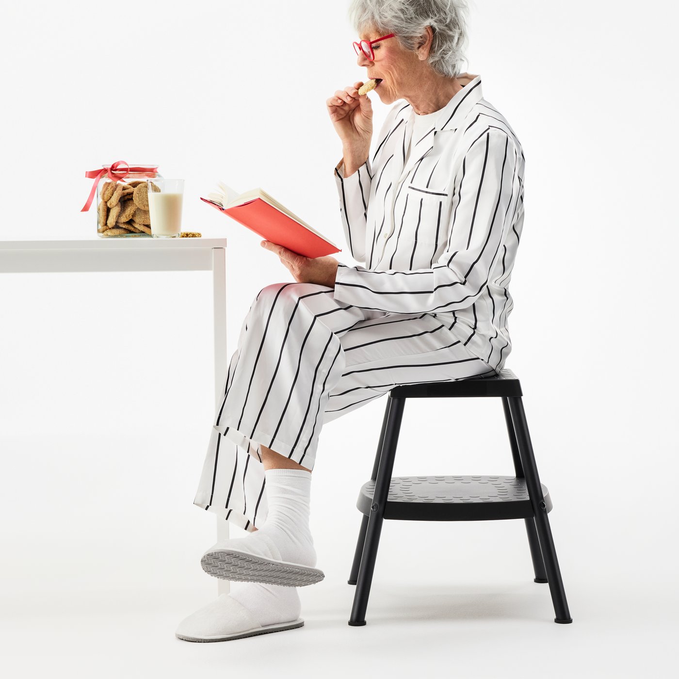 Femme en pyjama assise sur un marchepied HADDARP noir-gris et lisant un livre en mangeant des biscuits provenant d’un bocal à biscuits.