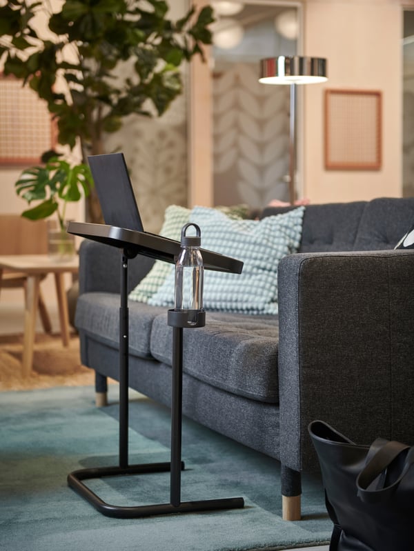Mysig lounge med mörkgrå LANDSKRONA soffa, turkosa kuddar och ett litet svart laptopbord med en vattenflaska.