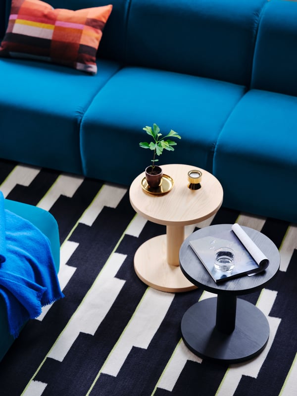 Two side tables sitting on a black/white and light green flatwoven STOCKHOLM 2025 rug next to a dark turquoise 3-seat sofa.