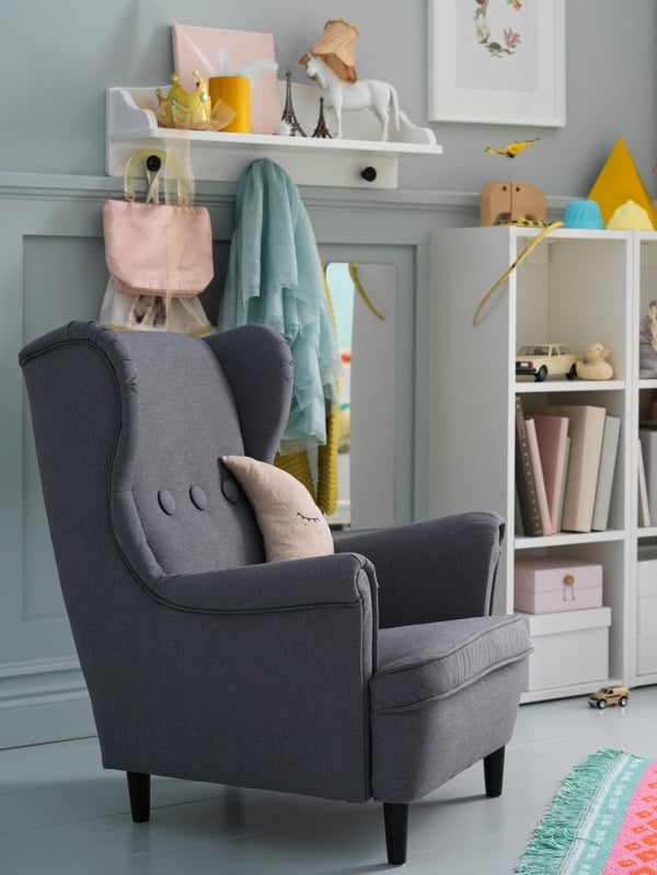 A grey STRANDMON children’s armchair stands next to a white SMÅGÖRA shelf unit that stores books, boxes and toys.