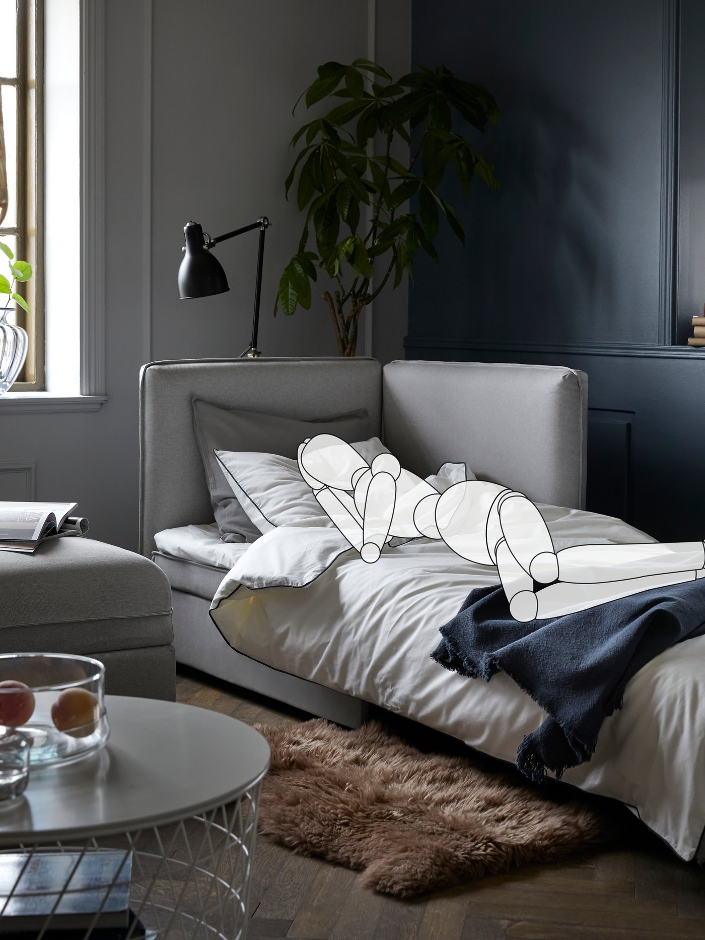 In a living room, a cartoon drawing of a person sleeps on a VALLENTUNA sofa-bed. A lamp and a plant are behind the sofa-bed.