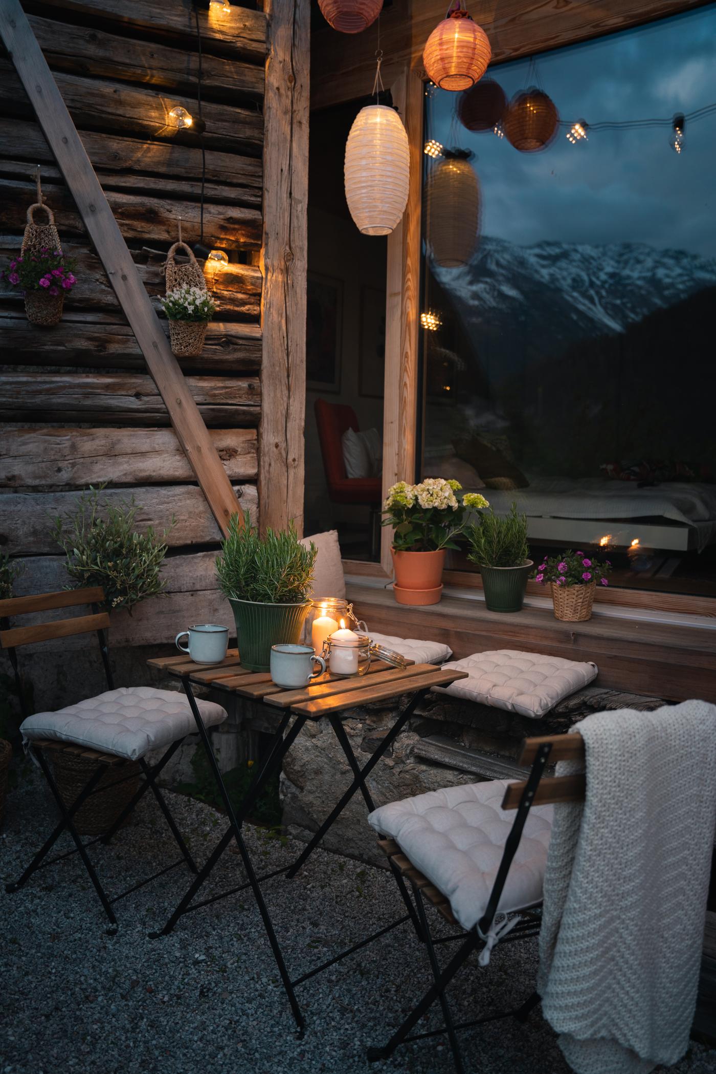 Gemütliche Terrasse bei Dämmerung mit Kerzen, Pflanzen und Laternen vor einem Chalet mit Alpenblick.