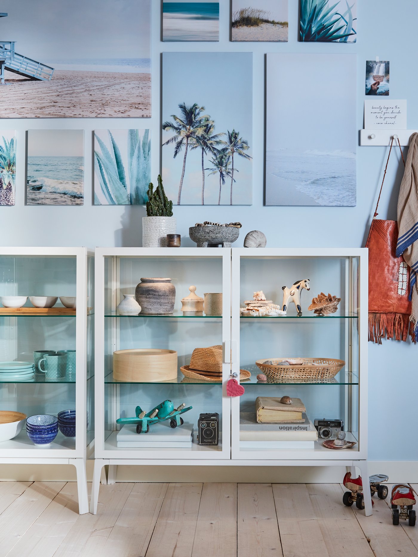Nine PJÄTTERYD beach pictures on a pale blue wall above a white MILSBO glass-door cabinet.
