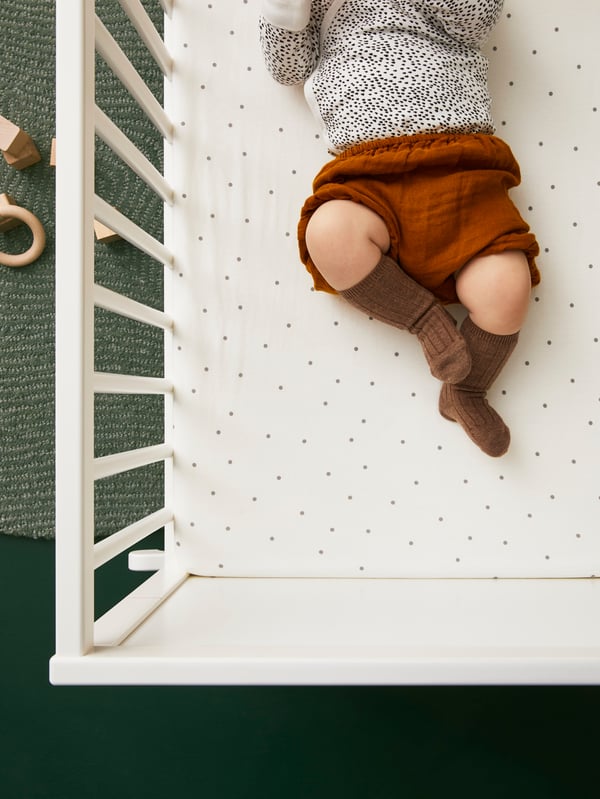 Un bébé portant short, chaussettes et combinaison dans un lit bébé couvert d’un drap housse LENAST à motifs à pois.