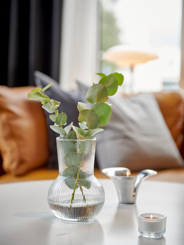 Detaljbild på ett vitt bord med en PÅDRAG glasvas med gröna kvistar och två värmeljushållare med tända ljus. I bakgrunden syns en ljusbrun IKEA STOCKHOLM skinnsoffa. 
