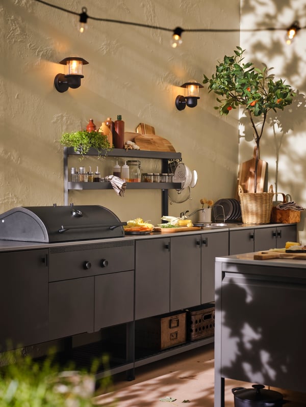 An outdoor wall with a BÅTSKÄR add-on unit with shelves over a BÅTSKÄR charcoal barbecue and unit for sink in dark grey.