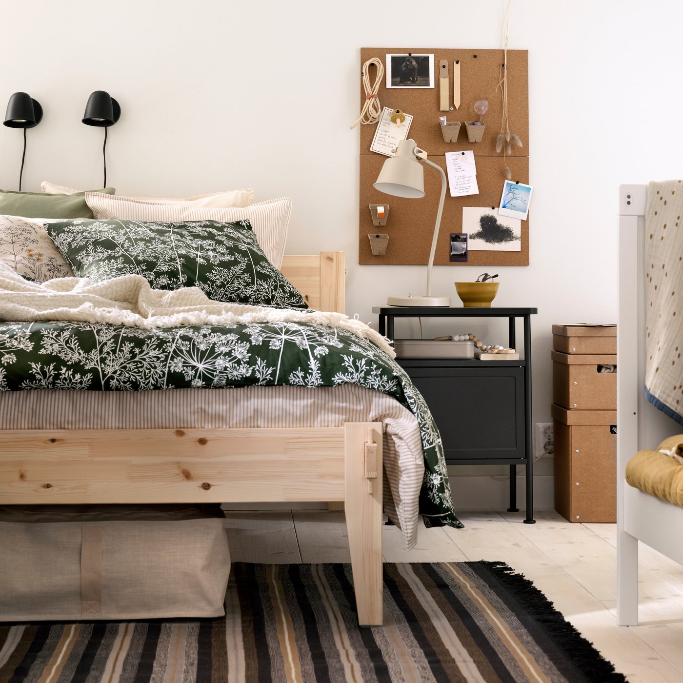 Un dormitorio con una cama TAXÅSKLINT de madera con un edredón de flores, una mesa de noche en gris oscuro y el borde de una cuna.