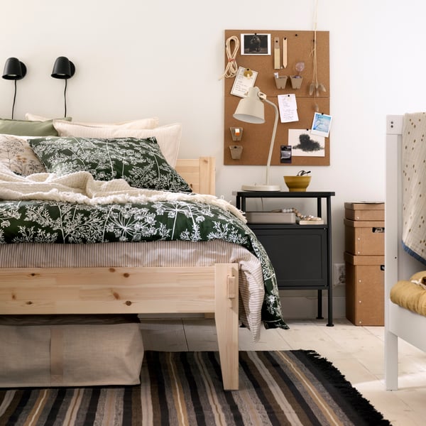 A bedroom featuring a wooden TAXÅSKLINT bed with a floral duvet, a dark grey nightstand and the edge of a child’s cot.