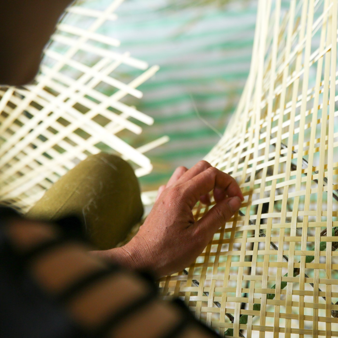 The hand of an Indian artisan hand-weaving a bamboo lattice from long, thin strands of bamboo.