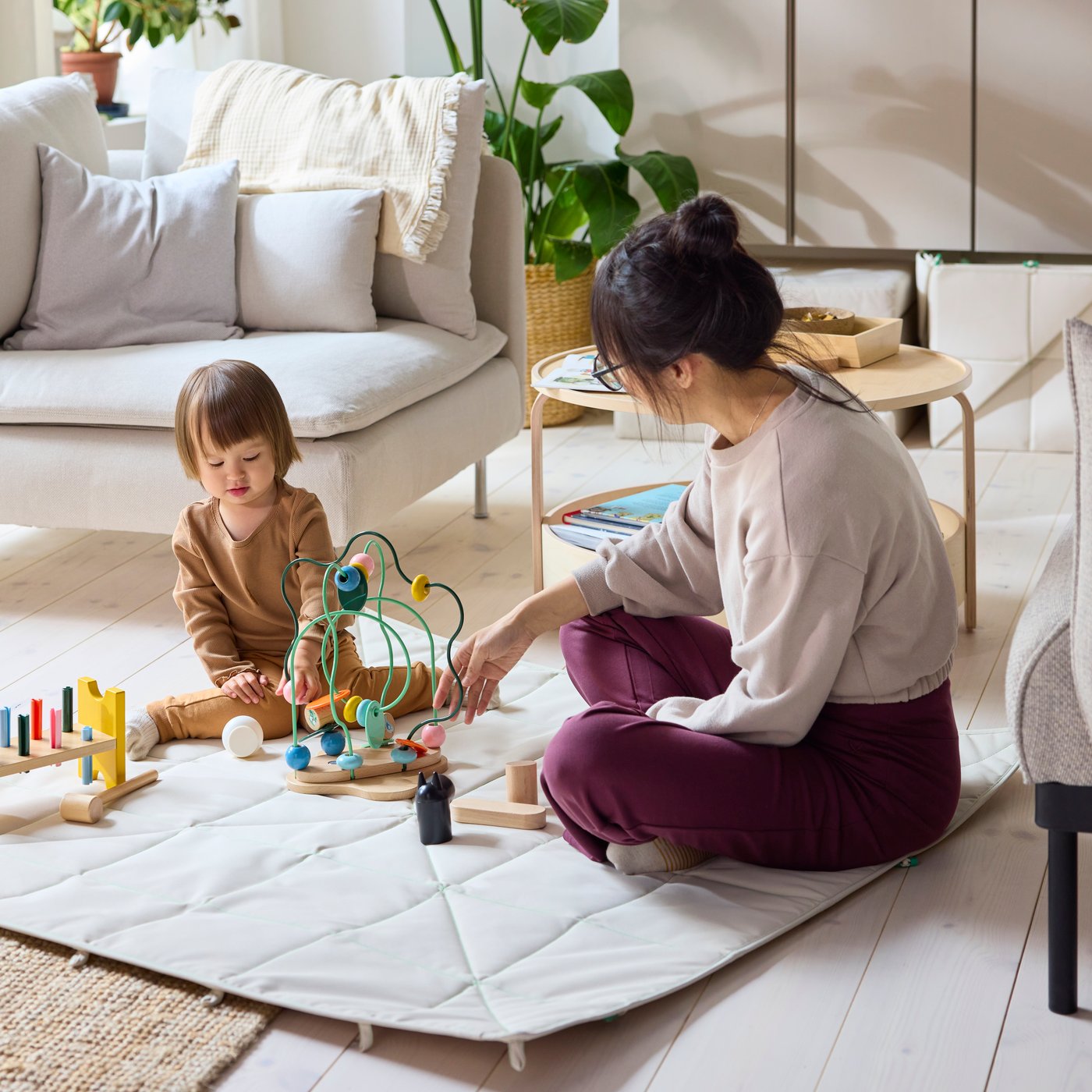 A beige GREJSIMOJS mat/foldable storage box pokes out from under a sofa in a modern living room. Soft toys are in the box.