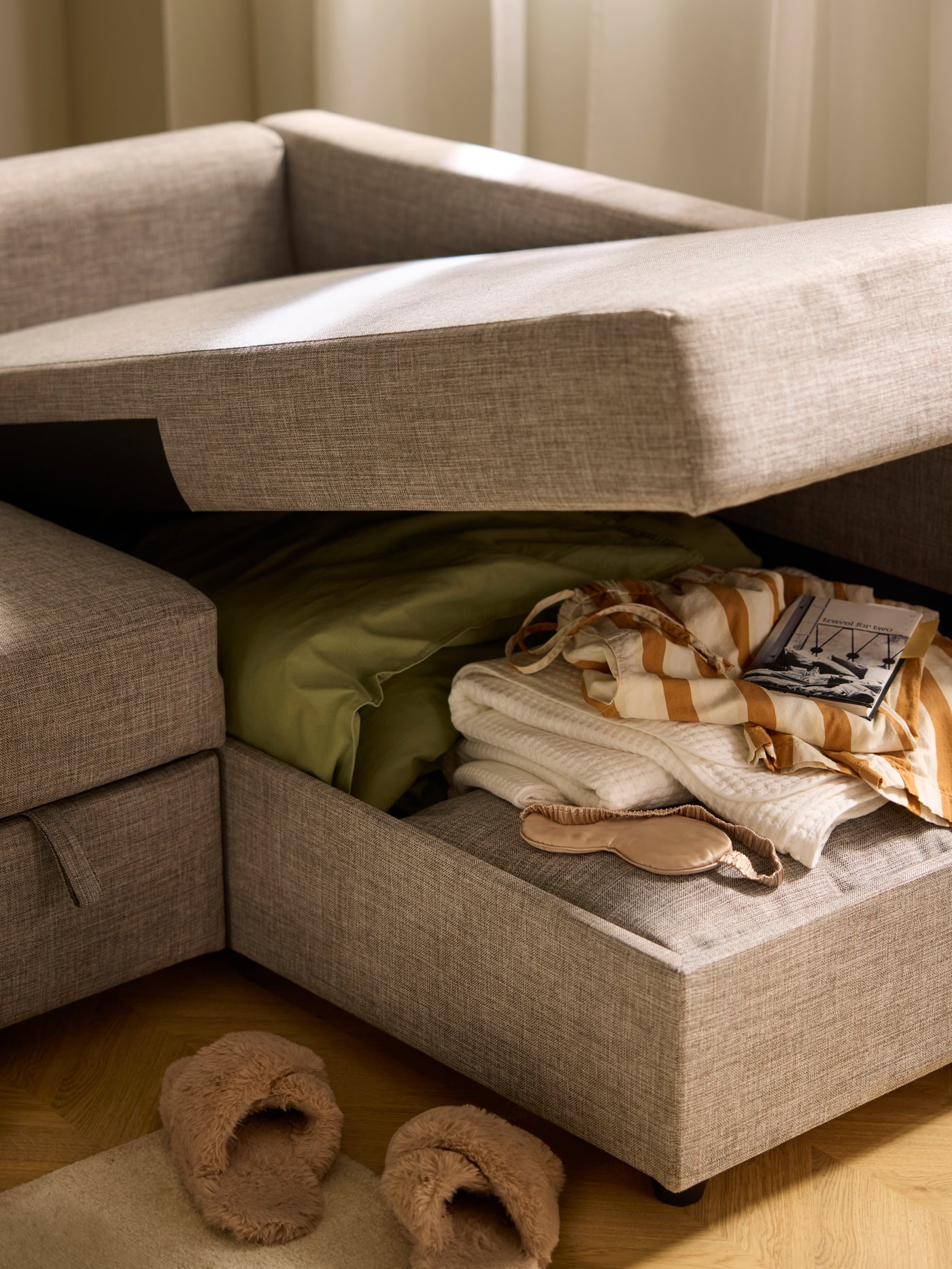 Slippers by the gaping chaise longue of a FRIHETEN sofa-bed, revealing bedding and textiles placed in the compartment within.