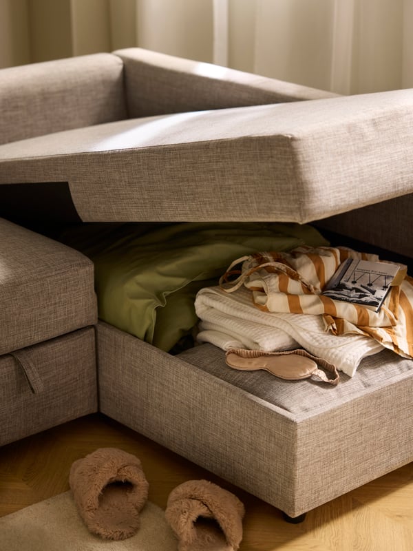 Papuče pored odškrinutog lenjivca FRIHETEN sofa ležaja u kom se vide posteljina i tekstil u unutrašnjem odeljku.