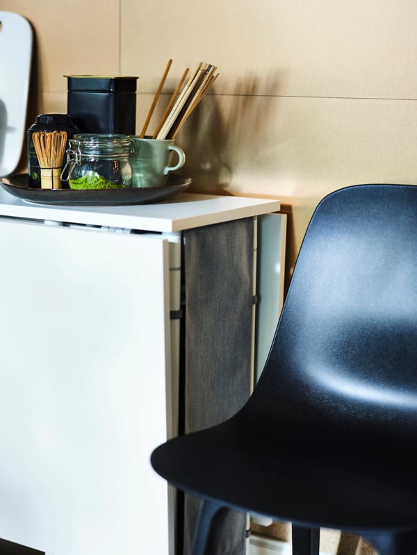 Ingeklapte KALLHÄLL klaptafel met opbergruimte in wit/lichtgrijs tegen een muur, naast een ODGER stoel in antraciet.