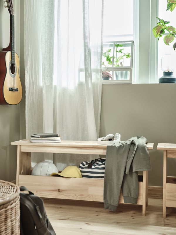 A PERJOHAN bench with storage in natural pine placed under a window with small plants. An acoustic guitar on the wall.