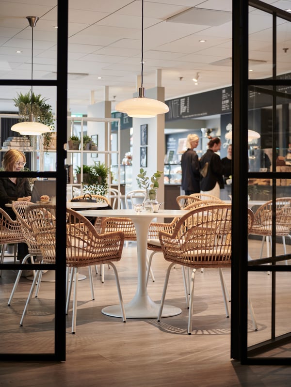 Genom glasdörrar med svart spröjs syns ett DOCKSTA matbord och NILSOVE stolar placerade i ett café. I bakgrunden syns en kassadisk med kunder och personal, 