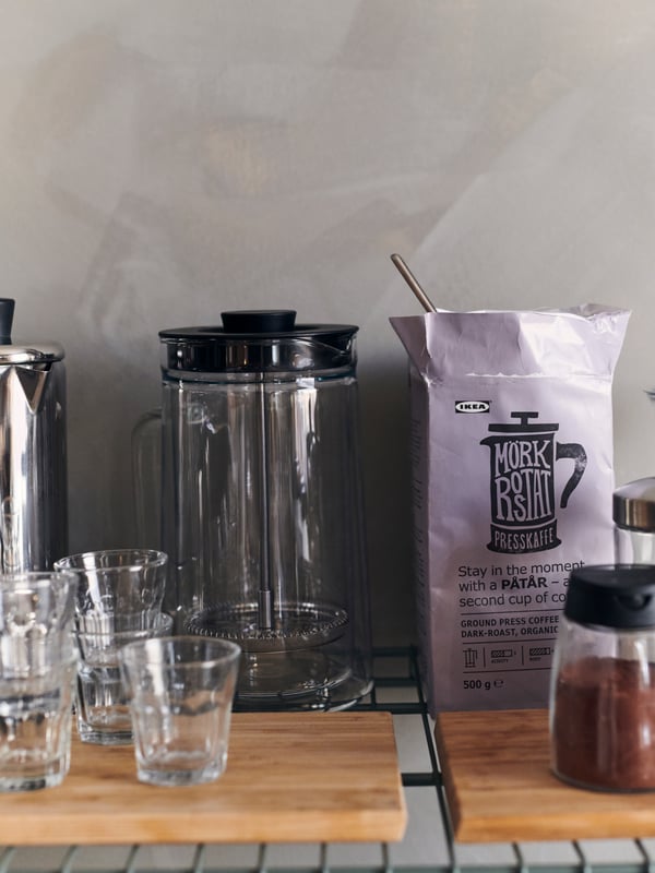 Une machine à café sur une planche à découper sur une étagère OMAR en métal, avec des verres POKAL autour et un sac de café.