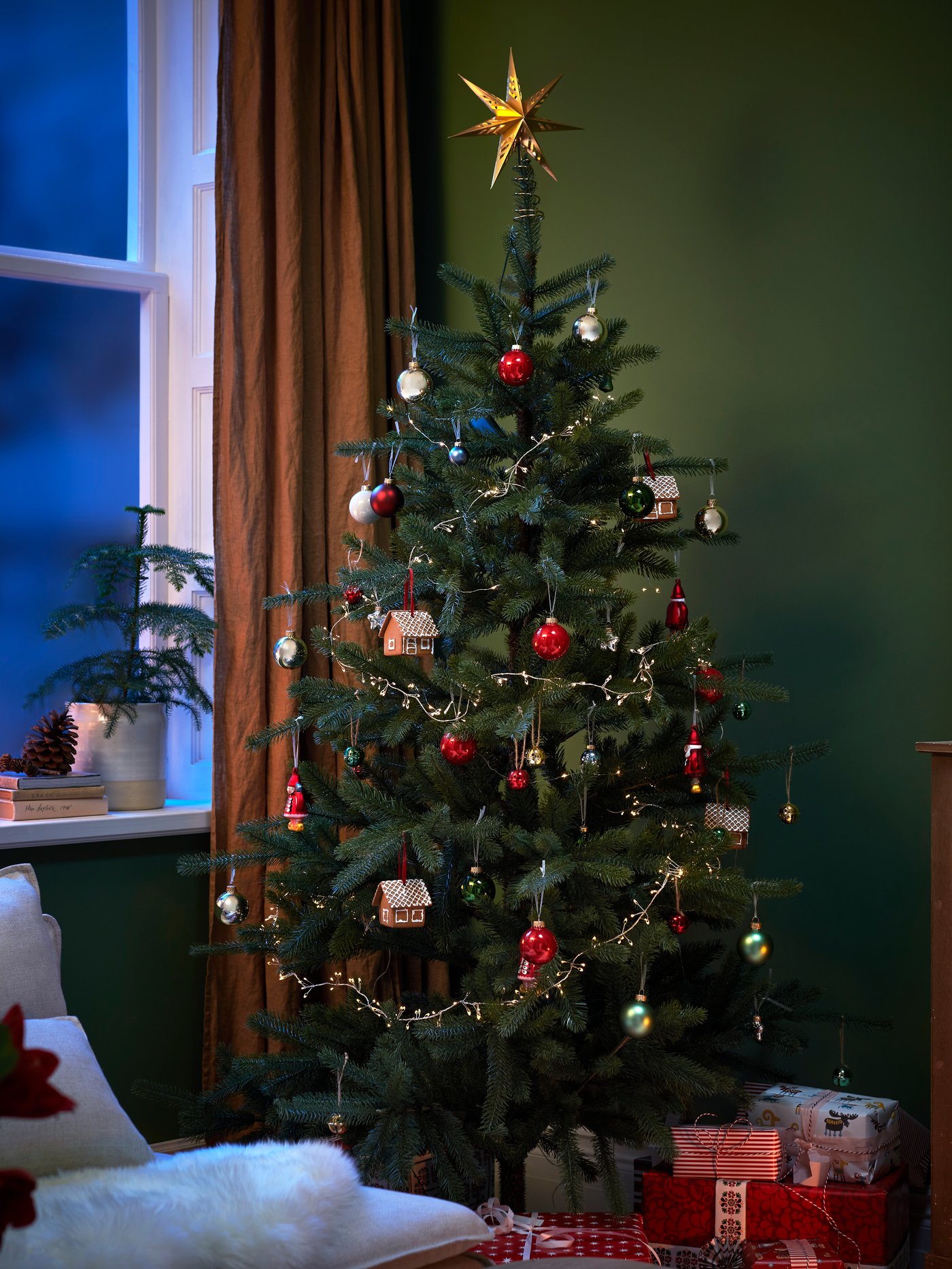 A Christmas tree near a window is decorated with VINTERFINT baubles and a STRÅLA LED tree topper star and lighting chain.