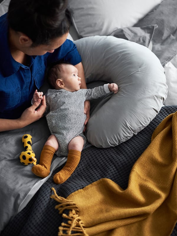 A woman sits on a bed resting her arm on a LEN nursing pillow and holding a baby. A yellow OMTÄNKSAM throw lies nearby.