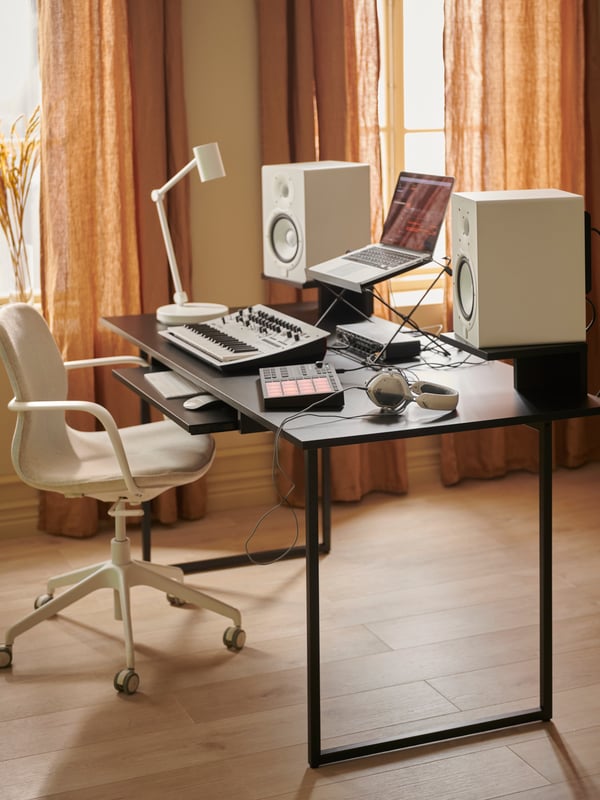A black OBEGRÄNSAD desk with white speakers, work lamp and swivel chair, laptop and music equipment in a light, beige room.