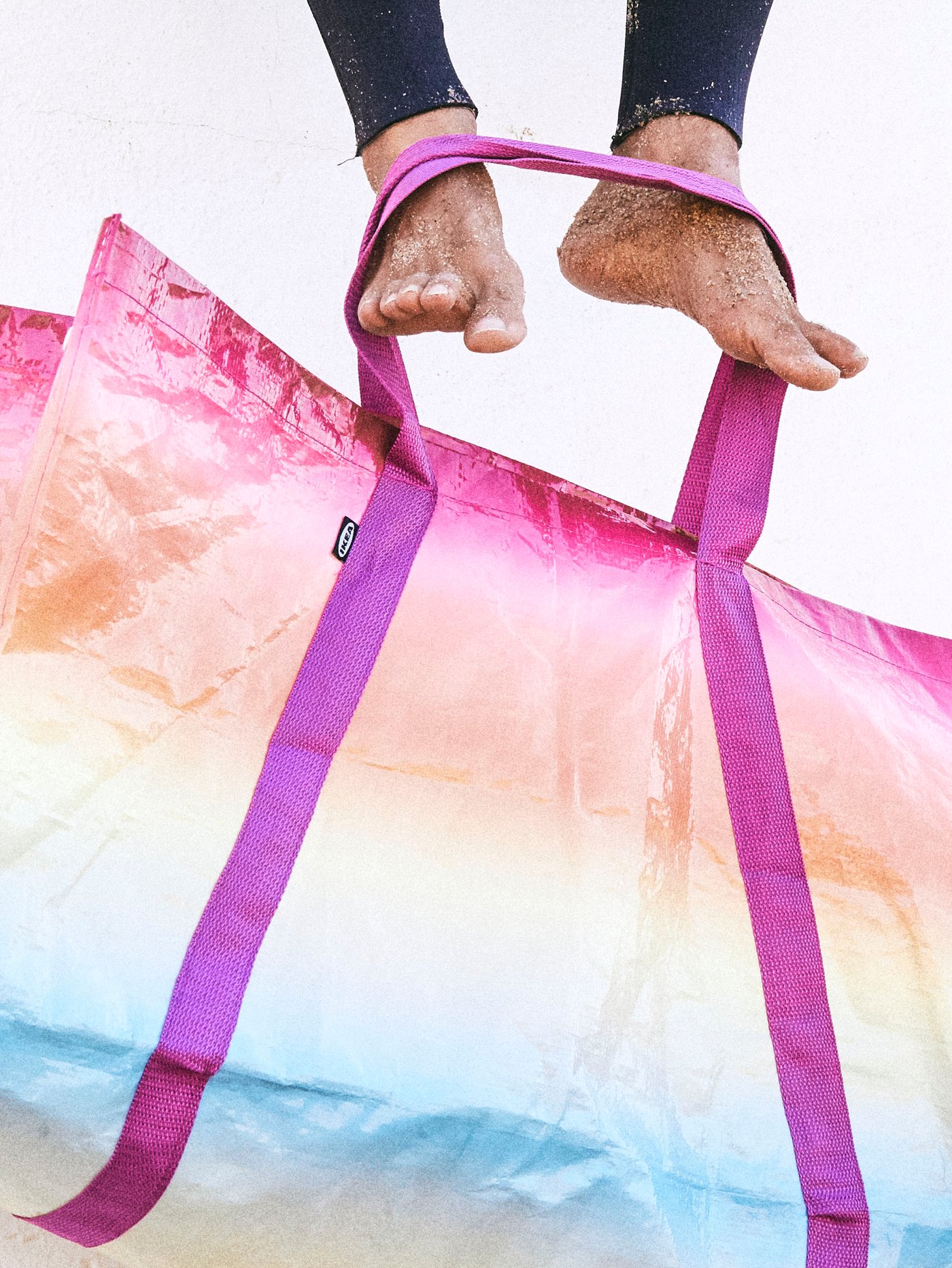 KÅSEBERGA bag made in a gradient of blues, yellows and pinks held by the feet of a person wearing a wetsuit.