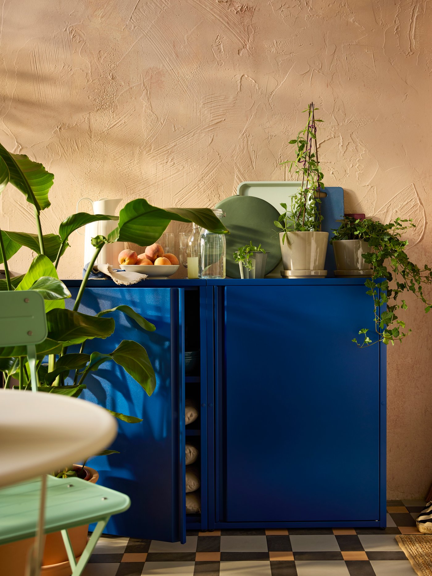 Accostati al muro di un balcone, due mobili SUNDSÖ blu scuro con piante, bicchieri e una caraffa termica TAGGÖGA bianco sporco.
